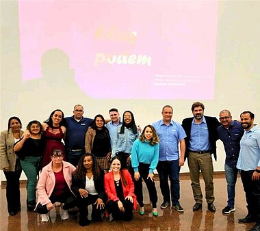 Group of diverse people on a stage. A pink presentation about Elas Podem in the background.